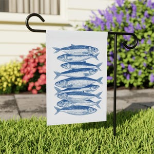 May include: A white garden flag featuring a blue illustration of sardines stacked vertically. The flag is displayed on a black metal stand in a grassy yard, with colorful flowers in the background.