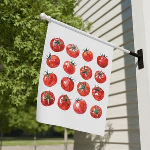 May include: A white outdoor flag featuring a pattern of red tomatoes with green stems. The tomatoes are arranged in a repeating pattern across the flag's surface. The flag is mounted on a white pole and is displayed outdoors.