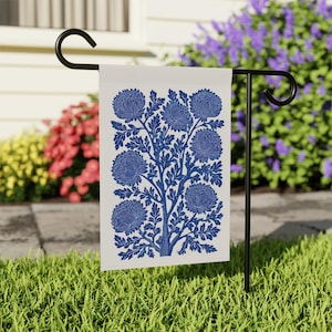 May include: A white garden flag with a blue floral design. The design features a stylized tree with large, round flowers and detailed leaves. The flag is displayed on a black metal stand in a grassy yard.