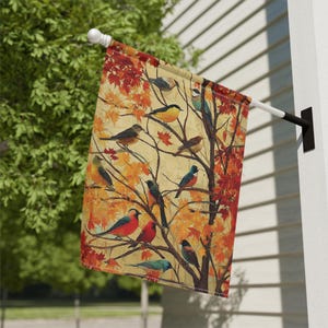 May include: A decorative outdoor flag featuring a detailed illustration of various birds perched on tree branches with autumn leaves. The flag has a cream-colored background with red, orange, and yellow leaves and birds in shades of blue, red, and yellow.