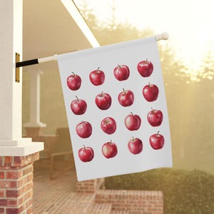 May include: A white flag with a pattern of red apples. The apples are evenly spaced across the flag's surface. The flag is hanging outside on a sunny day.