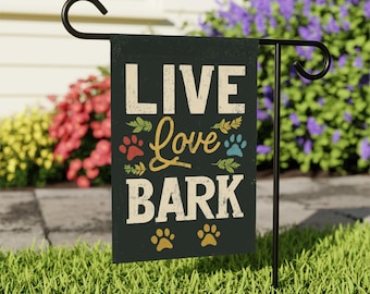 Peut inclure: Un drapeau de jardin noir avec les mots "LIVE Love BARK" en lettres blanches et dorées, avec des empreintes de pattes. Le drapeau est affiché sur un support en métal noir dans un jardin herbeux avec des fleurs colorées en arrière-plan.