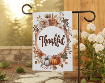May include: A white garden flag with the word "Thankful" in brown script, surrounded by a floral wreath with pumpkins and fall foliage. The flag is displayed on a black metal stand, perfect for autumn decor.