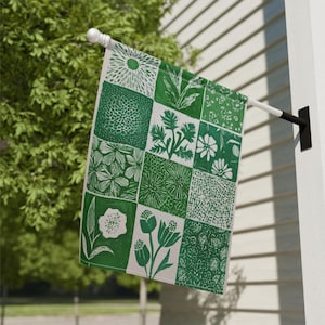 Può includere: Una bandiera da giardino verde e bianca con un design patchwork con varie illustrazioni botaniche. La bandiera è rettangolare e pende da un palo bianco, mostrando una collezione di motivi floreali e vegetali.