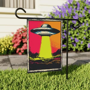 May include: A garden flag with a vintage-style image of a UFO emitting a yellow beam onto a landscape. The background fades from red to orange, framed by a black border. The flag is mounted on a black metal stand in a grassy garden.