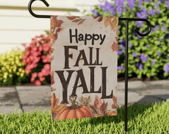 May include: A rectangular garden flag with the words "Happy Fall Y'all" in brown lettering. The flag features a pumpkin and autumn leaves in shades of orange, red, and yellow. The flag is displayed on a black metal stand in a grassy garden.