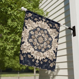 May include: A rectangular outdoor flag with a navy blue background and a beige and brown floral mandala design. The flag is mounted on a white pole and is hanging outside on a sunny day.