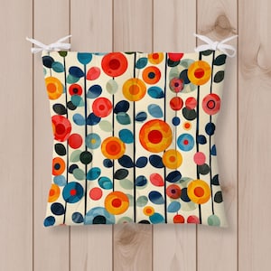 Puede incluir: Un cojín de asiento cuadrado con fondo crema y un patrón repetitivo de líneas verticales negras y motivos florales circulares coloridos en tonos rojo, naranja, amarillo y azul. El cojín incluye lazos de tela blanca en las esquinas superiores.