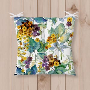 Peut inclure: Un coussin de siège carré avec un fond blanc et un motif floral à l'aquarelle dans des tons de jaune, violet et vert sarcelle. Le coussin comprend quatre attaches en tissu blanc aux coins pour le fixer au cadre d'une chaise.