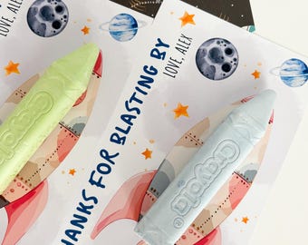 May include: Two Crayola crayons, one light green and one light blue, rest on cards with rocket ship illustrations. The cards have the text "THANKS FOR BLASTING BY LOVE, ALEX" and feature planets and stars. A space-themed background is visible.