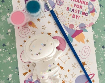 May include: A space-themed craft kit with a white astronaut figurine, paint pots in pink and blue, a paintbrush, and a circular card that says "THANKS FOR BLASTING BY!". The background is a light green with star patterns.