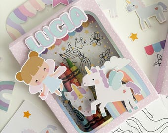 May include: A pink crayon box with the name "Lucia" in light blue letters. The box features a window with crayons and unicorn illustrations. Additional unicorn and fairy stickers, rainbows, and stars are scattered around the box.