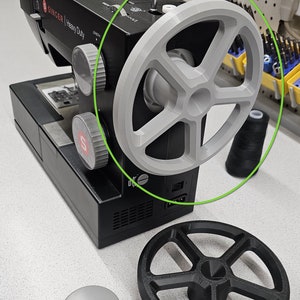 Könnte beinhalten: Eine schwarze Singer Heavy Duty Nähmaschine mit einem grauen Handrad. Ein schwarzes Handrad und eine graue Spulenkapsel befinden sich vor der Maschine. Eine Spule schwarzen Fadens befindet sich rechts.