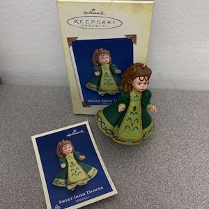 May include: A Hallmark Keepsake Ornament featuring a doll in a green dress with gold accents. The doll has brown hair and is labeled "Sweet Irish Dancer". The ornament comes with a box and a card with the same image.