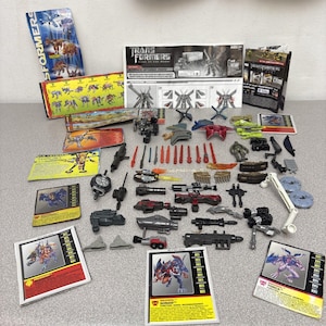 May include: A collection of Transformers toys and accessories, including weapons, cards, and instruction manuals. The toys are in various colors, including gray, red, and green. The cards feature character illustrations and names.