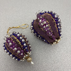 May include: Two egg-shaped ornaments covered in purple fabric, decorated with vertical rows of dark blue beads, white faux pearls, and purple sequins. Each ornament features intricate gold wire trim and a gold-toned metal cap at the base.