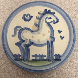 Puede incluir: Un plato redondo de cerámica con base color crema y un diseño azul pintado a mano. El centro presenta un caballo estilizado con manchas de color marrón claro, de pie detrás de una pequeña valla. El borde está decorado con una gruesa franja azul.