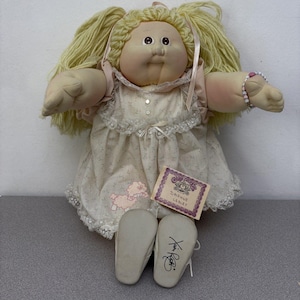 Puede incluir: Una muñeca de tela con cabello de lana rubia en coletas, vestida con un vestido color crema con estampado floral pequeño, ribete de encaje y un gráfico de cordero rosa. La muñeca tiene una pulsera de cuentas y una etiqueta Daphne Lebley, firma en la suela.