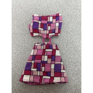May include: A two-piece doll outfit in shades of pink, purple, and white. The top is a halter style, and the skirt is a mini-skirt. The fabric has a geometric pattern of squares and rectangles.