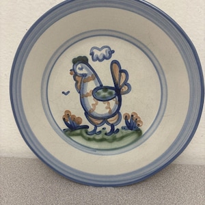 Puede incluir: Un cuenco redondo de cerámica con base color crema y bandas azules en el borde. El centro presenta una ilustración pintada a mano de un gallo en azul, beige y verde, de pie sobre un trozo de hierba con pequeñas flores y una nube.