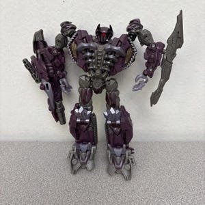 May include: A purple and gray action figure of a robot with articulated joints. The figure has a detailed design with various weapons and armor components. The robot's head features a red eye and a unique helmet design. The figure is posed with its arms raised.