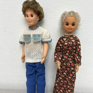 May include: Two vintage doll figures. One doll wears a white knit shirt with blue trim and blue pockets, paired with blue pants. The other doll is dressed in a long floral print dress with a burgundy background and pink flowers.