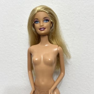 Puede incluir: Una muñeca rubia con ojos azules y una expresión neutra. La muñeca tiene el pelo largo y liso y no lleva ropa. El tono de piel de la muñeca es claro y el fondo es blanco liso.