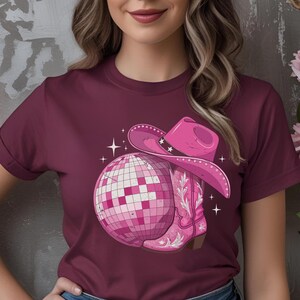 K&ouml;nnte beinhalten: Burgunderfarbenes T-Shirt mit einem rosa Motiv einer Discokugel, einem Cowboyhut und einem Cowboy-Stiefel. Der Hut hat Sternakzente. Der Stiefel hat ein Blumenmuster. Wei&szlig;e Glitzer umgeben die Grafik.