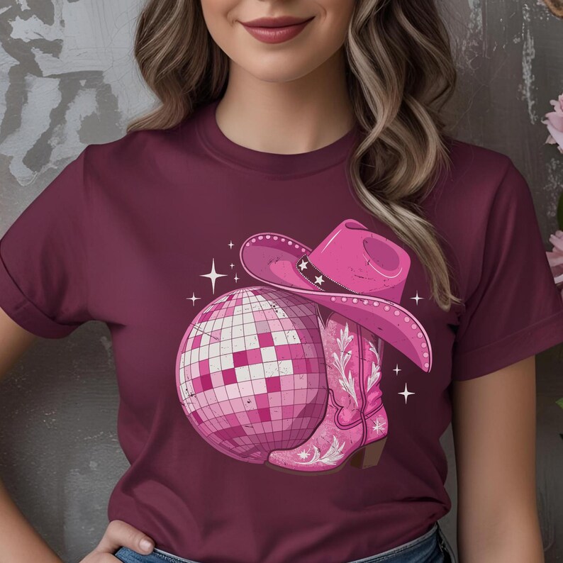 K&ouml;nnte beinhalten: Burgunderfarbenes T-Shirt mit einem rosa Motiv einer Discokugel, einem Cowboyhut und einem Cowboy-Stiefel. Der Hut hat Sternakzente. Der Stiefel hat ein Blumenmuster. Wei&szlig;e Glitzer umgeben die Grafik.