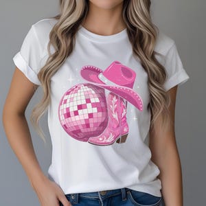 K&ouml;nnte beinhalten: Wei&szlig;es T-Shirt mit einem Design aus einer rosa Discokugel, Cowboyhut und Cowboy-Stiefel. Der Stiefel hat ein Blumenmuster. Das Design ist in Rosa- und Wei&szlig;t&ouml;nen gehalten, mit Sternakzenten.