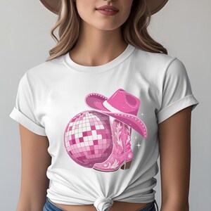 K&ouml;nnte beinhalten: Wei&szlig;es T-Shirt mit einem pinkfarbenen Discokugel-, Cowboyhut- und Cowboy-Stiefel-Motiv. Der Stiefel hat ein florales Design. Der Hut und der Stiefel sind dunkler rosa als die Discokugel. Wei&szlig;e Sternakzente vervollst&auml;ndigen das Design.