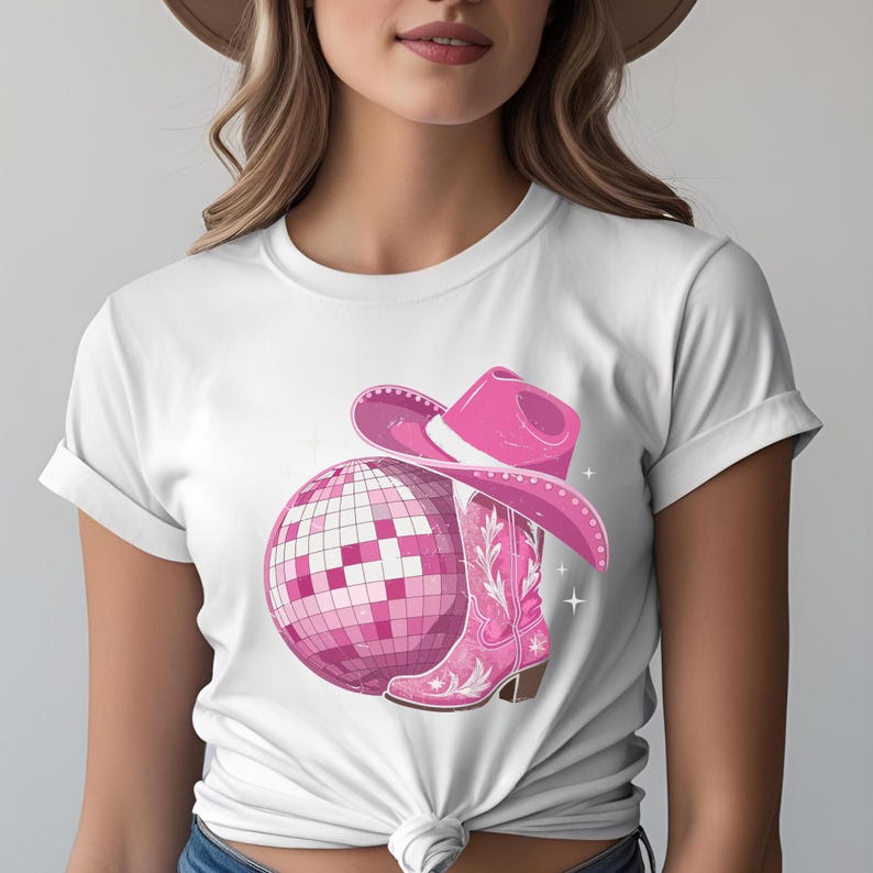 K&ouml;nnte beinhalten: Wei&szlig;es T-Shirt mit einem pinkfarbenen Discokugel-, Cowboyhut- und Cowboy-Stiefel-Motiv. Der Stiefel hat ein florales Design. Der Hut und der Stiefel sind dunkler rosa als die Discokugel. Wei&szlig;e Sternakzente vervollst&auml;ndigen das Design.