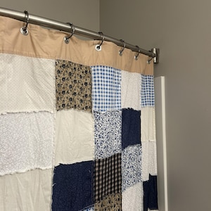 May include: A patchwork shower curtain with a tan top and a variety of fabric patterns in white, blue, and brown. The curtain hangs from a silver rod with metal rings, creating a rustic, country-style bathroom decor.