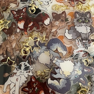 May include: A collection of cat-themed keychains with gold-colored star-shaped clasps. The keychains feature various cat designs in shades of brown, white, gray, and orange. Some cats have green accents. The keychains are made of clear acrylic.