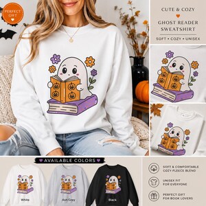 Könnte beinhalten: Ein weißes Sweatshirt mit Rundhalsausschnitt und einer Grafik eines Cartoon-Geistes, der ein orangefarbenes Buch mit Kürbisgesichtern liest, auf einem lila Buch sitzt und von orangefarbenen und lila Blumen umgeben ist.