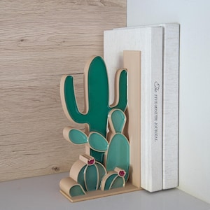 May include: A decorative cactus-shaped bookend in shades of green and beige. The bookend is holding two books, one of which has the text "The Five-Minute Journal" printed on the spine. The bookend is made of a glossy material.