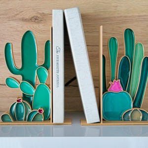 May include: Cactus-shaped bookends in shades of green, teal, and pink, with gold accents. The bookends are holding two books, one with the text "The FIVE-MINUTE JOURNAL". The bookends are on a white surface with a wood-grain background.