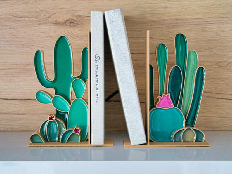 May include: Cactus-shaped bookends in shades of green, teal, and pink, with gold accents. The bookends are holding two books, one with the text "The FIVE-MINUTE JOURNAL". The bookends are on a white surface with a wood-grain background.