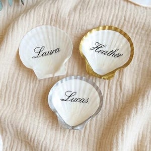 Peut inclure: Trois coquilles Saint-Jacques blanches portant les prénoms Laura, Heather et Lucas en écriture cursive noire. Une coquille a un bord doré, une autre un bord argenté et la troisième est unie. Une quatrième coquille affiche le nom Veamar.