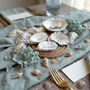 May include: A collection of oyster shells used as place cards, featuring the names VeaMar, Heidi, Eddy, and Roland written in black script. Some shells have gold or silver painted edges, arranged on a circular jute rope base with small seashells.