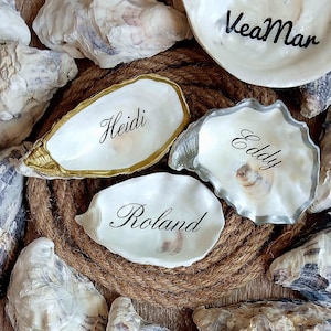 Op de afbeelding: Vier witte oesterschelpen met de namen Heidi, Eddy, Roland en VeaMar in zwarte kalligrafie. Eén schelp heeft een goudkleurige rand, één een zilverkleurige rand en twee zijn effen. De schelpen liggen op een opgerold bruin jute touw.