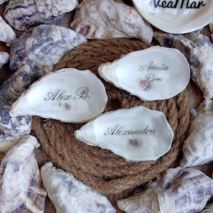 Op de afbeelding: Drie witte oesterschelpen met gladde, parelmoerachtige binnenkanten, elk voorzien van een naam in een zwart cursief lettertype. De namen zijn Alex B., Amelia Rossi en Alexandra. De schelpen zijn gerangschikt op een ronde, opgerolde bruine touwbasis.