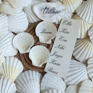 May include: A collection of white scallop shells arranged around a circular jute rope. Two shells feature the names Veamar and Laura in black script. A rectangular white card lists the names Ellie, Kim, Eva, Niki, Jaya, Kaia, and Beau in black script.