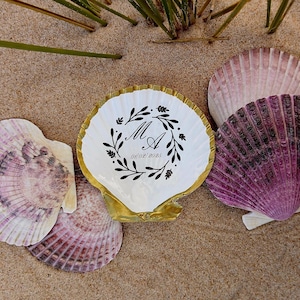 Può includere: Una conchiglia di capasanta bianca con bordo dipinto in oro, caratterizzata da un design a ghirlanda botanica nera che circonda le lettere MA e la data 01/02/2025, circondata da quattro conchiglie naturali viola e bianche su una superficie sabbiosa.