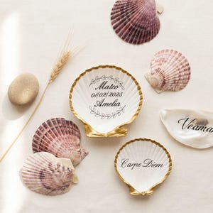 Può includere: Due conchiglie di capasanta bianche con bordi dipinti in oro, una con il testo Mateo 01/02/2025 Amelia e un motivo a foglie, l'altra con il testo Carpe Diem. Sono circondate da conchiglie naturali, una pietra e una spiga di grano.