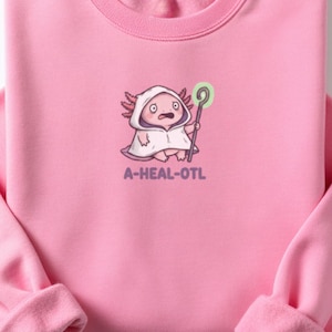 May include: Pink sweatshirt featuring a cartoon axolotl character wearing a hooded cloak and holding a staff with a glowing green orb. The text "A-HEAL-OTL" is printed below the character. The sweatshirt has a crew neck and long sleeves.