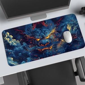 Puede incluir: Alfombrilla de escritorio rectangular con una ilustración detallada de la cara de un dragón entre nubes arremolinadas azules y naranjas. Un ratón blanco descansa sobre la alfombrilla, junto a un cuaderno rojo y una cámara. También se ven un portátil y una pequeña planta.