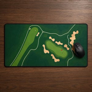 May include: A rectangular desk mat featuring a top-down graphic illustration of a golf course layout with green fairways, tan sand bunkers, and white contour lines. A black computer mouse rests on the right side of the mat surface.