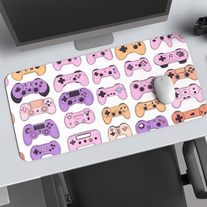 May include: A white desk mat featuring a pattern of colorful game controllers in shades of purple, pink, and orange. A computer mouse rests on the mat, alongside a laptop, monitor, camera, and a red notebook.