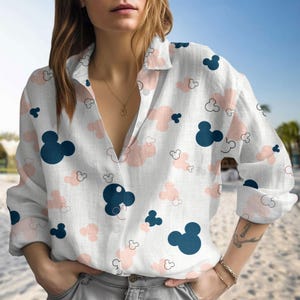 May include: White button-down shirt with a pattern of navy blue and pink Mickey Mouse silhouettes and cloud shapes. The long-sleeved shirt has a V-neck collar.